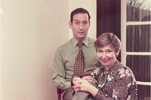 As Joy's husband sits in a chair, Joy sits beside him on the floor with her hands clasped on his knee.